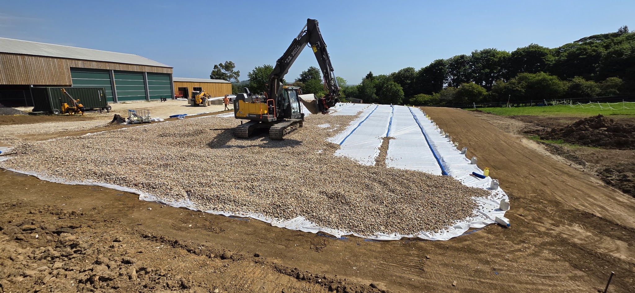 Stone base going down over geotextile beside farm buildings