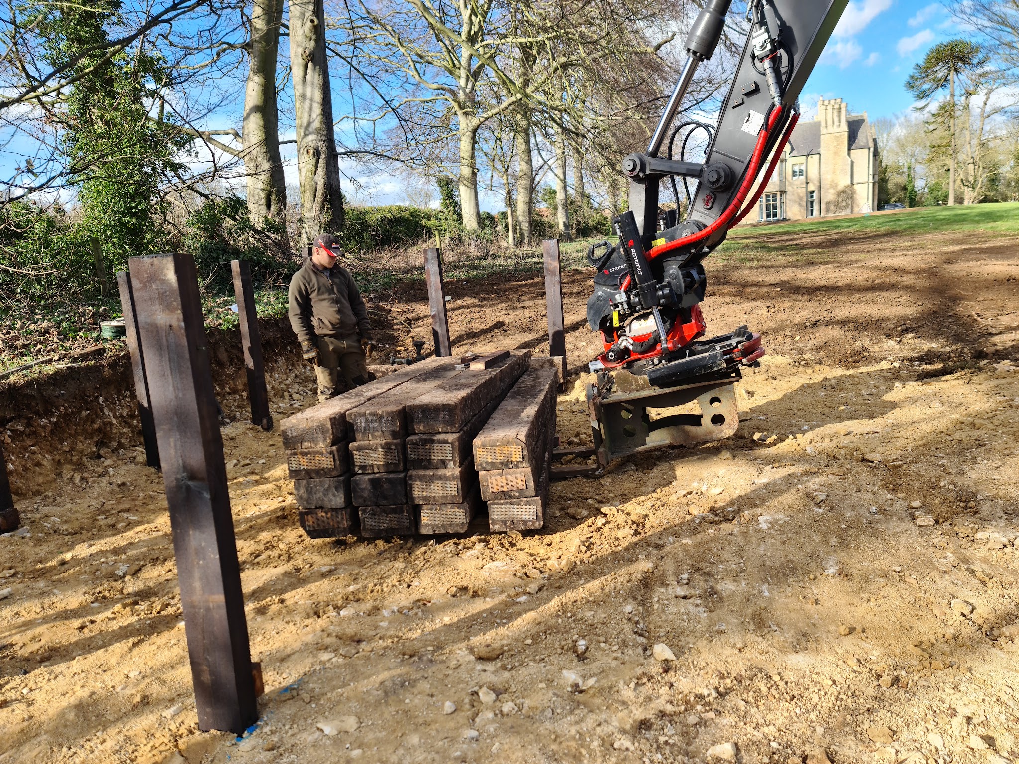 Timber sleepers being placed by a tiltrotator in front of a manor house